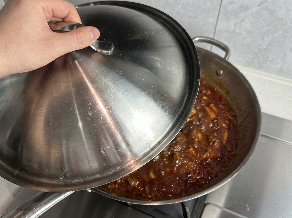 A hand placing a stainless steel lid onto a large wok-style pan filled with bubbling red meat sauce.