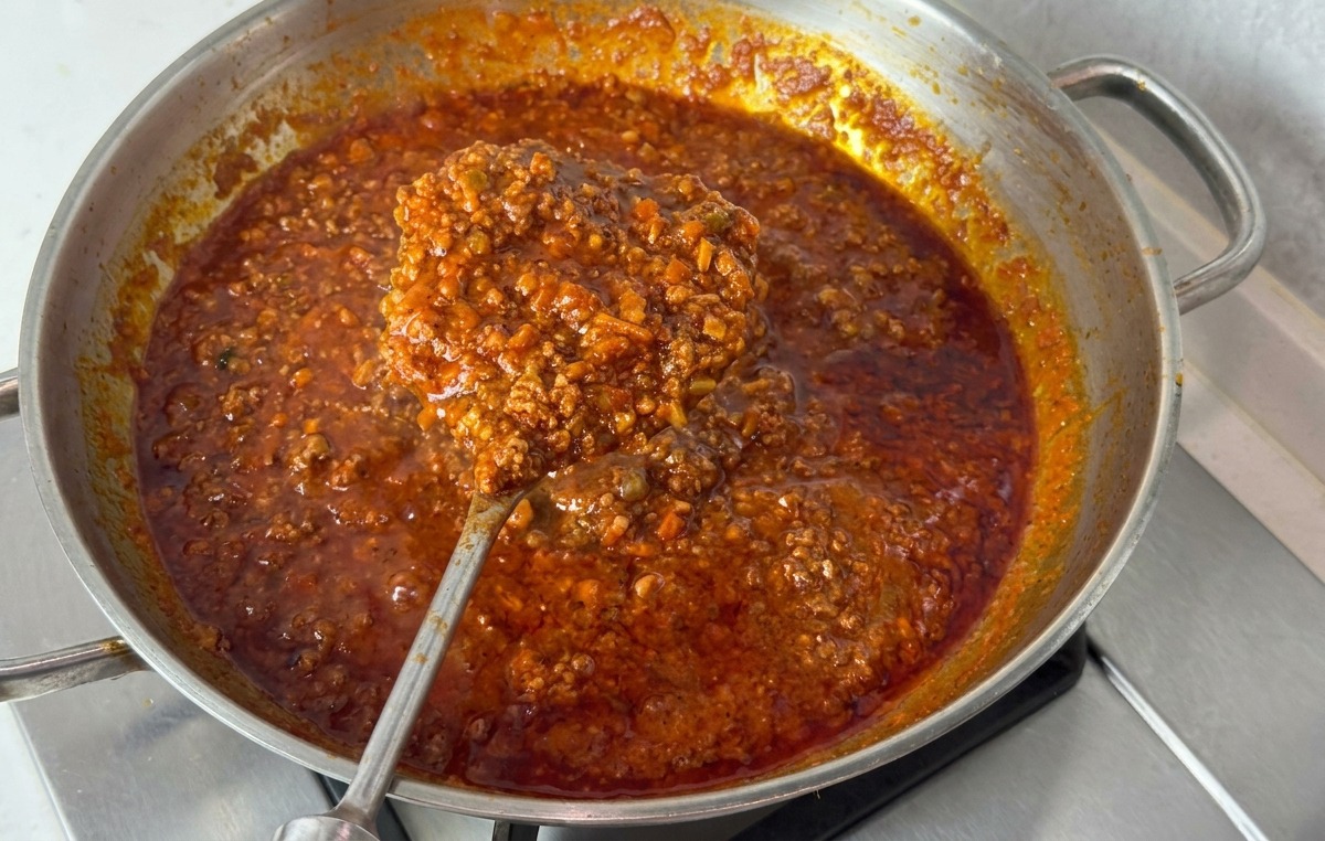 A stainless steel pan filled with thick, dark red Bolognese-style sauce being lifted with a large metal spoon to show its viscous, bubbling consistency.