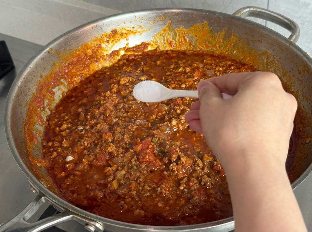 A hand holding a small white plastic spoon filled with fine white salt over a pan of bubbling red meat sauce.