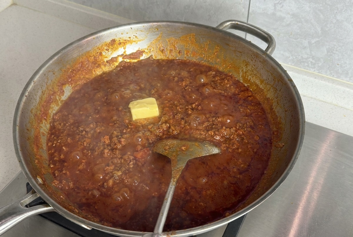 A square yellow block of butter resting on the surface of a simmering red tomato meat sauce in a large silver pan.