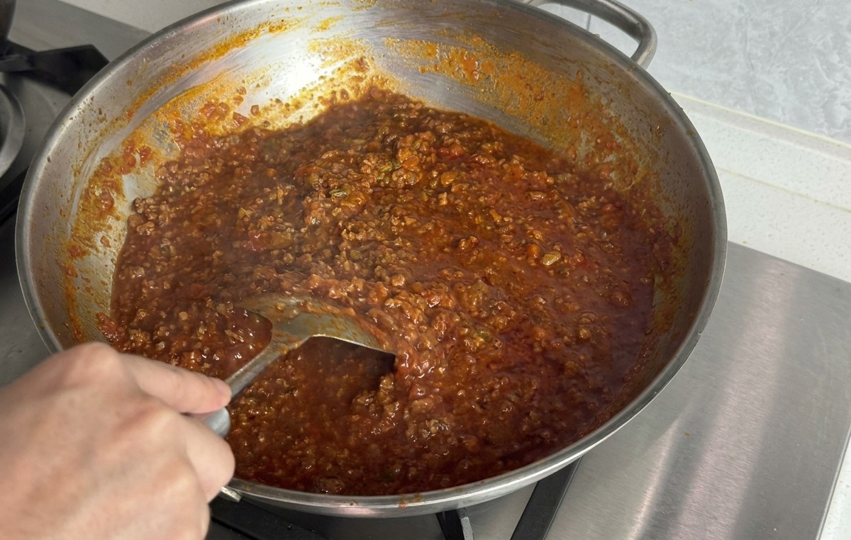 A hand using a metal spatula to stir a melting pat of butter into a thick, dark red meat sauce in a stainless steel pan.