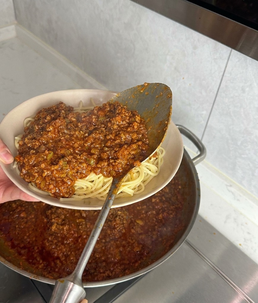A large metal ladle pouring thick, chunky red meat sauce onto a bed of plain cooked spaghetti noodles in a beige bowl.