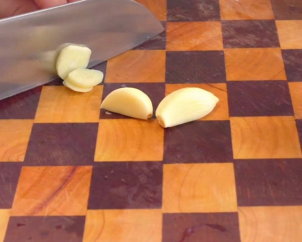A hand using a chefs knife to cut peeled garlic cloves into thin slices on a wooden board.