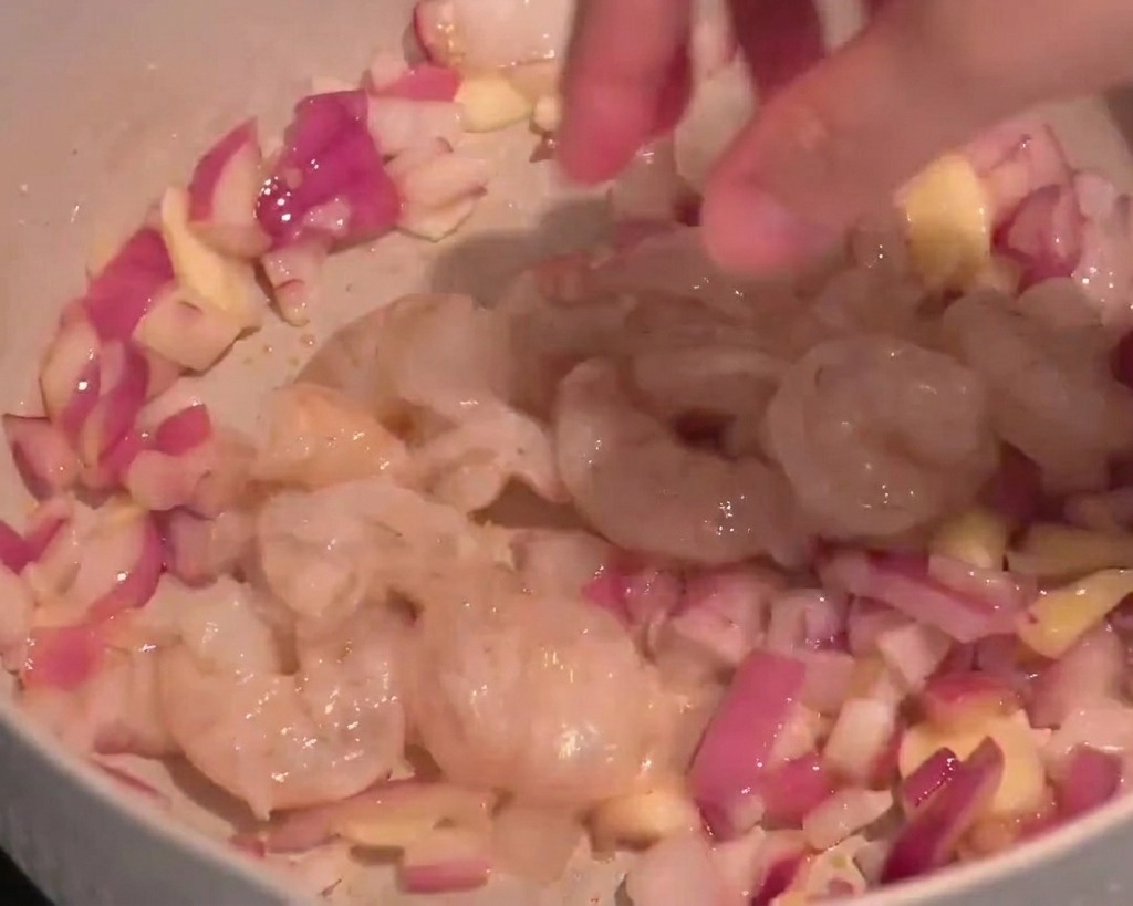 Translucent raw shrimp being added to a pan with chopped red onions and sliced garlic.