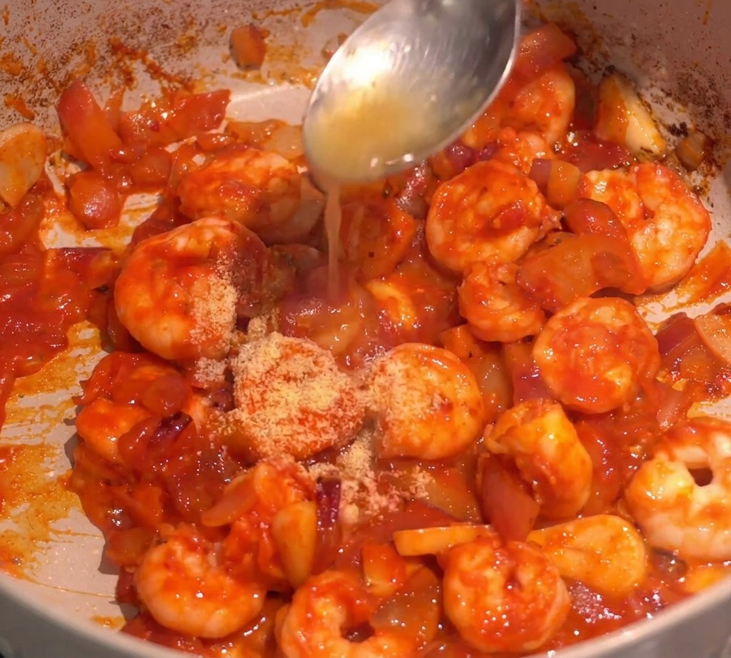 Spices being added to a pan of shrimp in a red chili sauce, with liquid being poured from a spoon.