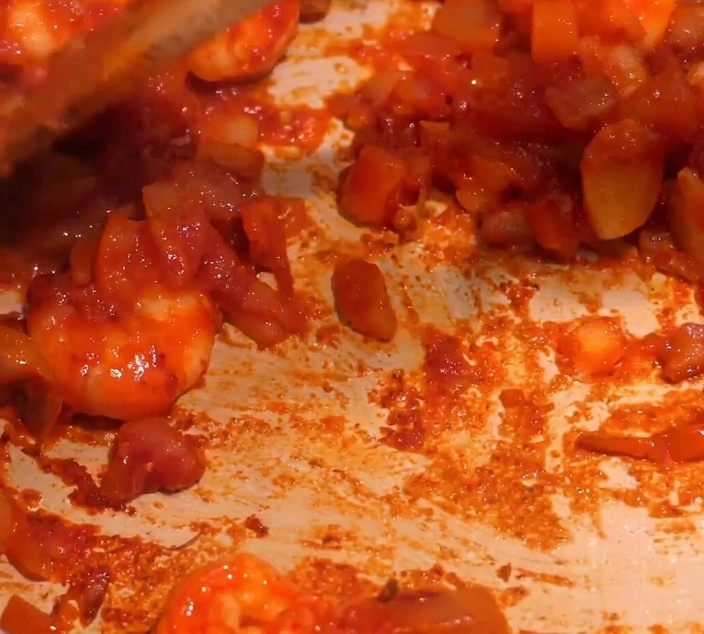 A thick, red shrimp sauce being stirred in a pan as liquid is added to create a simmer.