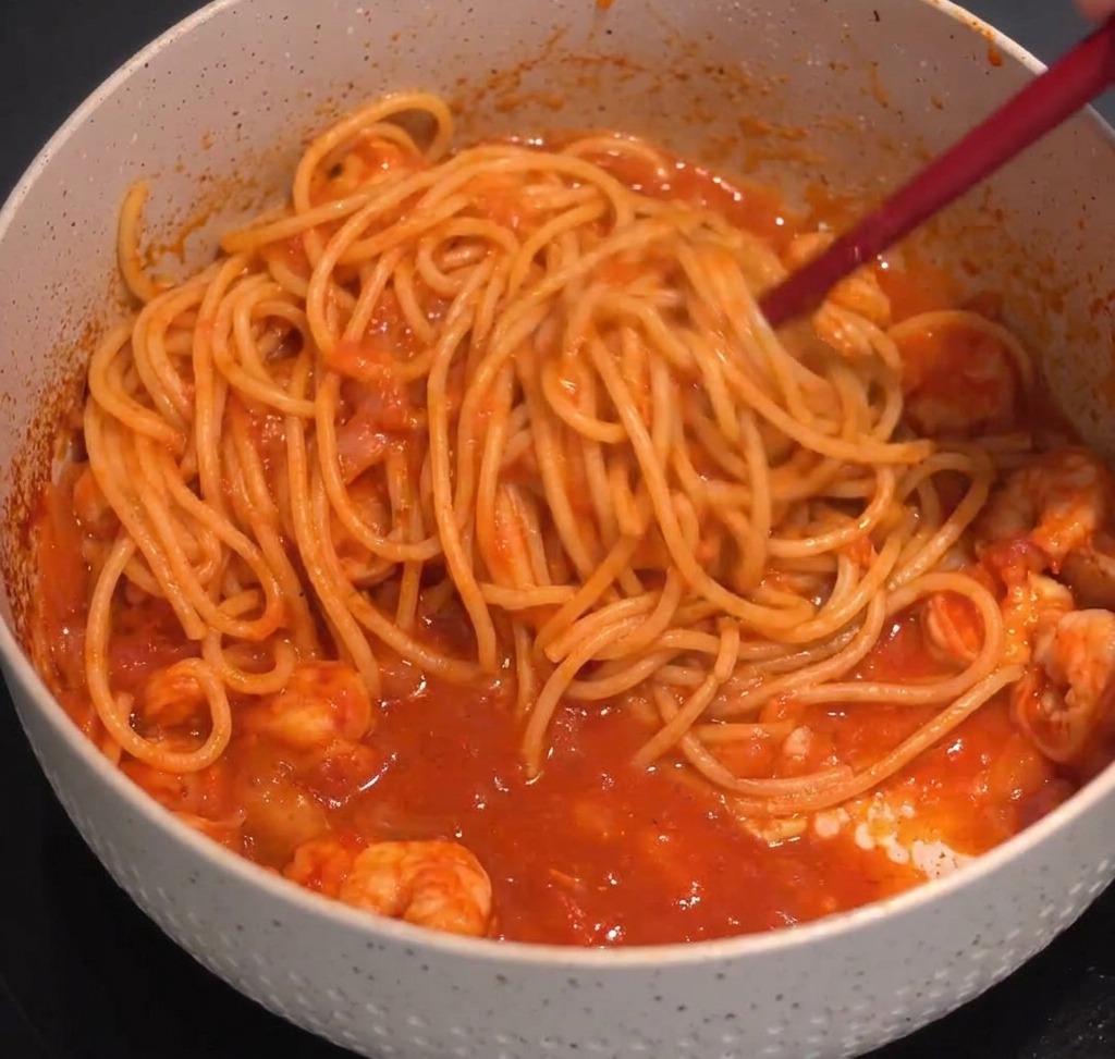 Spaghetti being tossed with red shrimp sauce using kitchen tongs in a speckled pot.