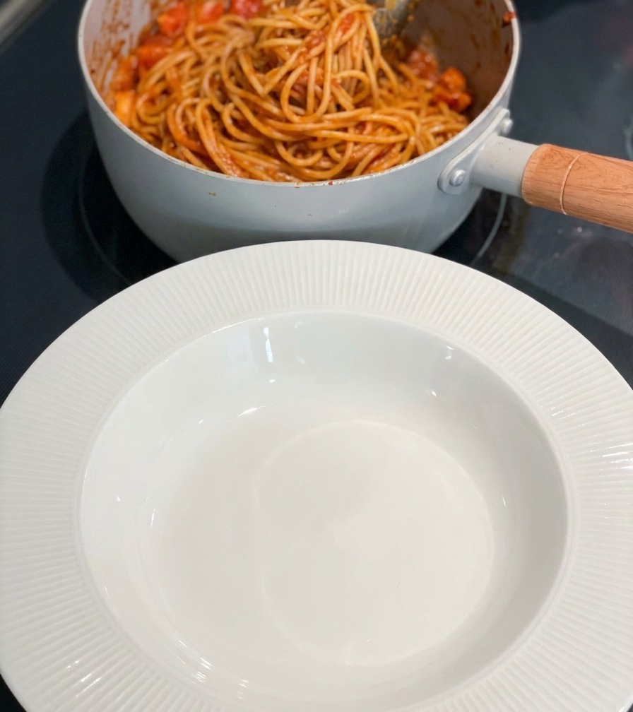 A pot of finished spicy shrimp pasta next to an empty white rimmed bowl on a cooktop.