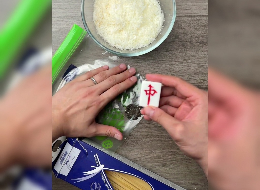 A hand using the flat side of a mahjong tile to crush black peppercorns inside a plastic bag on a wooden table.