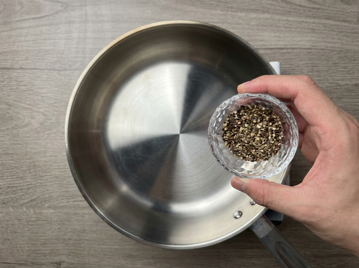 Coarsely crushed black pepper being poured from a small glass bowl into a dry preheated stainless steel pan.