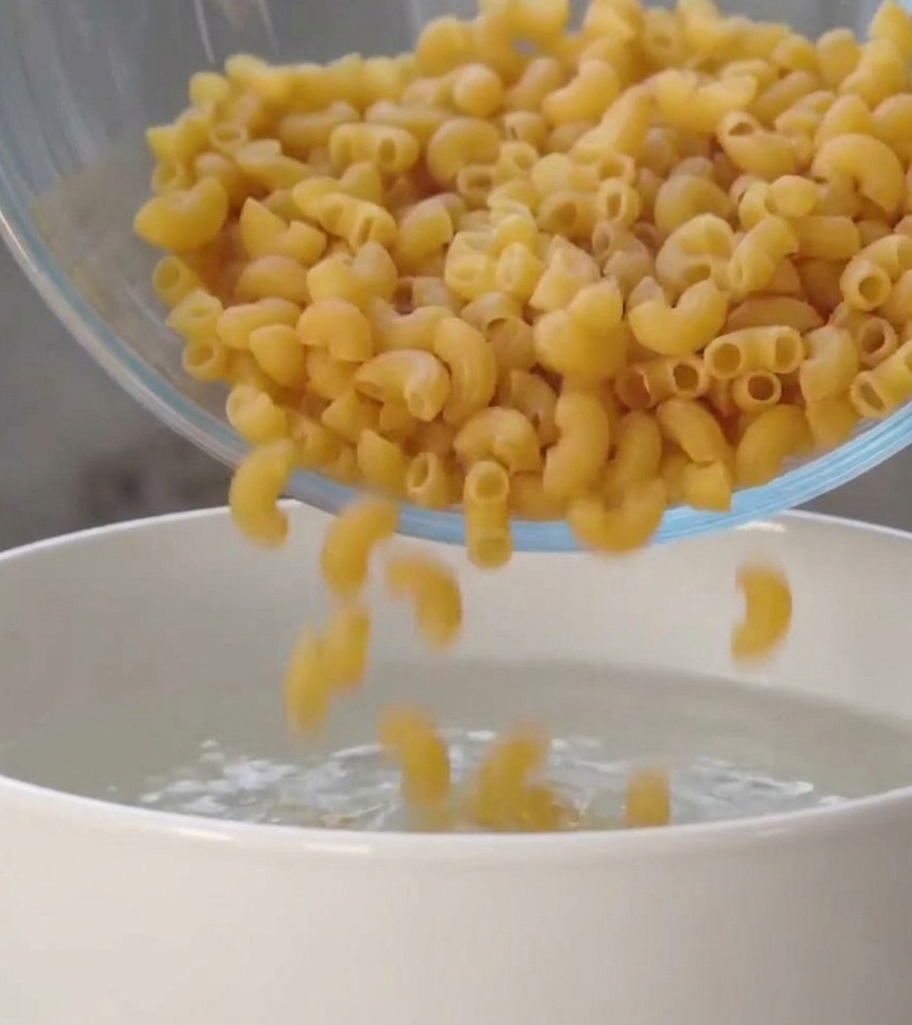 Dry spiral pasta pieces falling from a clear glass bowl into a white pot filled with clear water