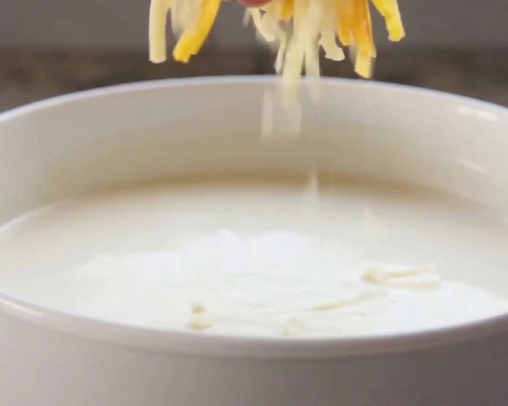 A hand dropping shredded orange and white cheese into a bowl of warm white cream sauce