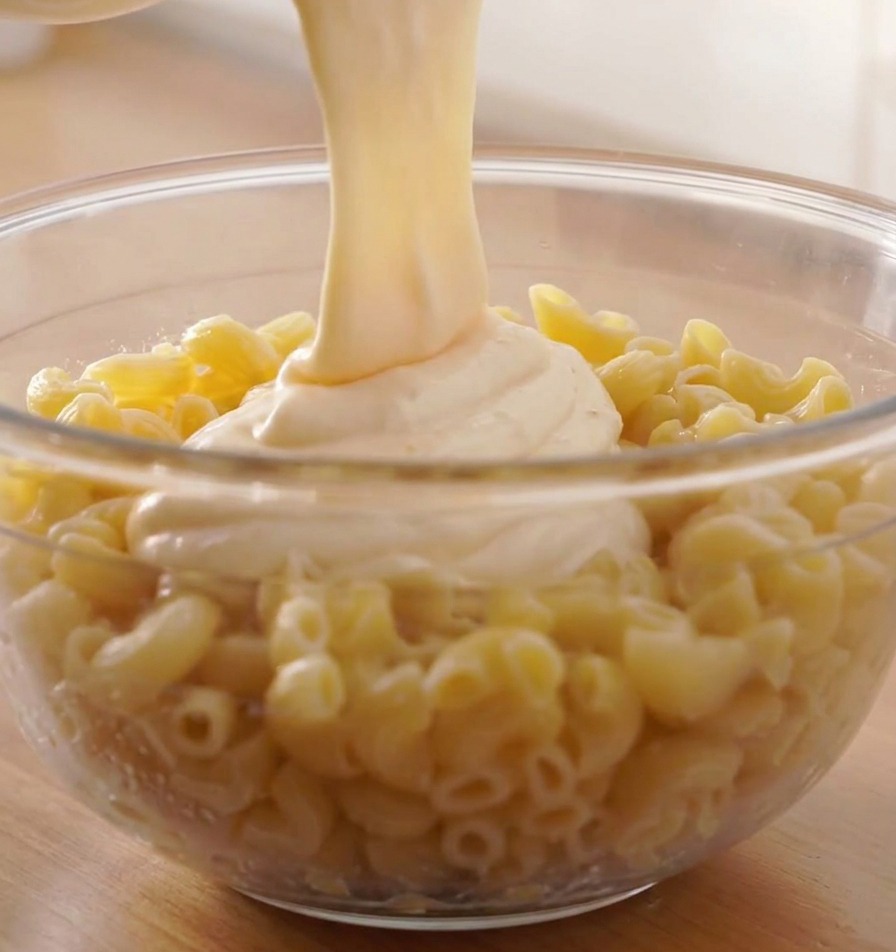 A thick stream of melted white cheese sauce being poured over a large bowl of cooked spiral pasta