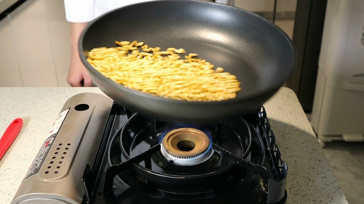 Pine nuts being tossed in a black non-stick frying pan over a blue gas flame.