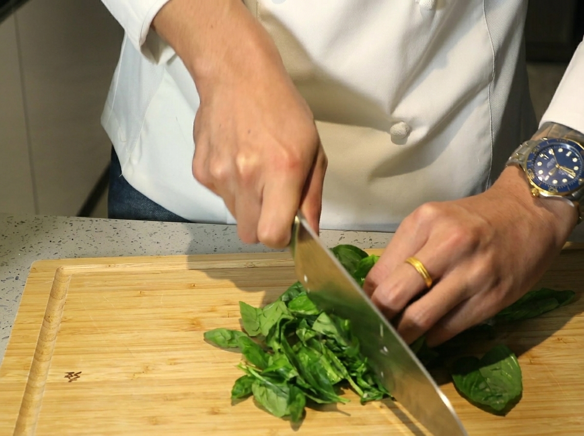 Hands using a large knife to chop a pile of fresh green basil leaves on a wooden board.