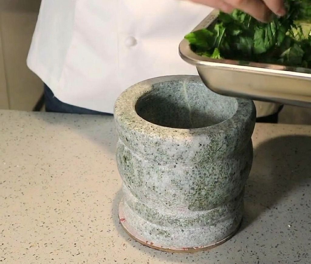 A person adding chopped fresh basil leaves from a metal tray into a heavy granite mortar.