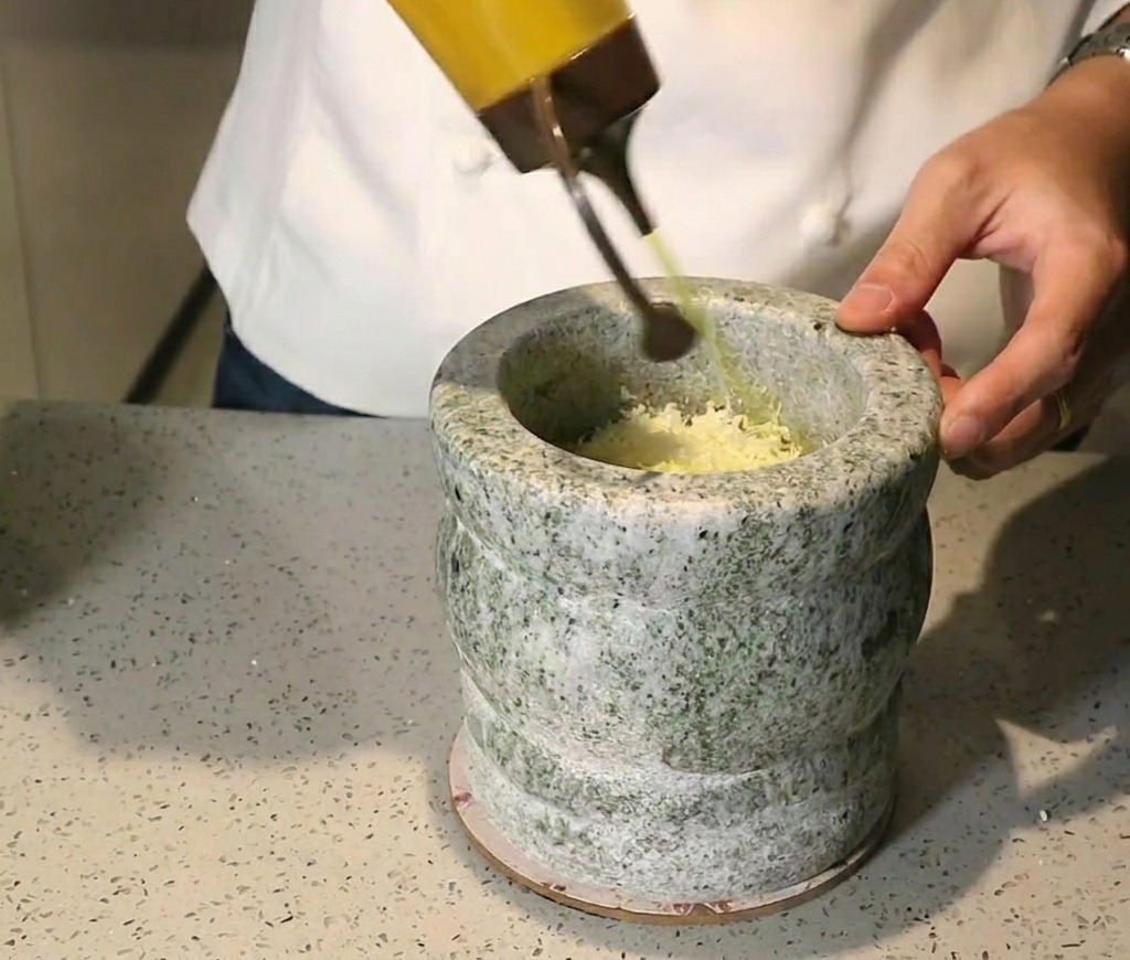 Golden olive oil being poured from a squeeze bottle into a stone mortar filled with crushed green herbs and nuts.