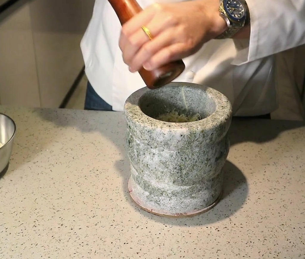 A wooden pepper mill being used to grind peppercorns over the ingredients inside a stone mortar.