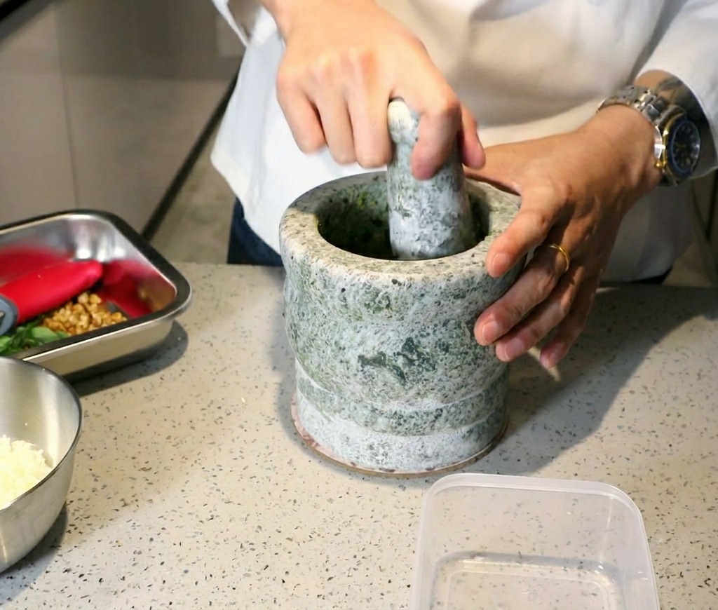 A chef using a stone mortar and pestle to crush fresh basil and garlic into a thick green pesto paste.