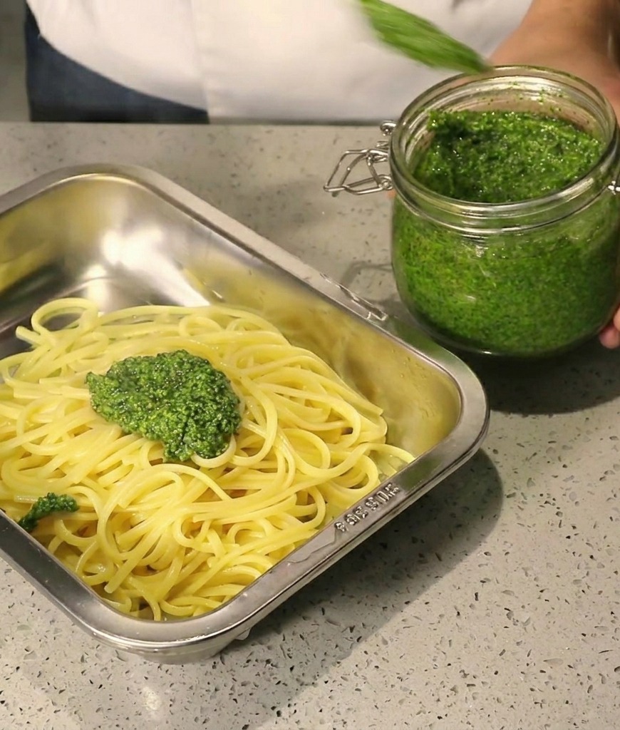 Thick green pesto being spooned from a glass jar onto hot spaghetti in a stainless steel tray.
