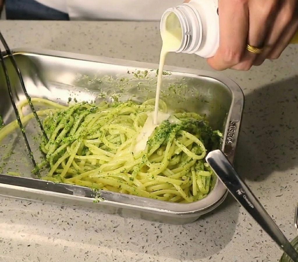 A stream of creamy white sauce being poured onto pasta topped with green pesto.