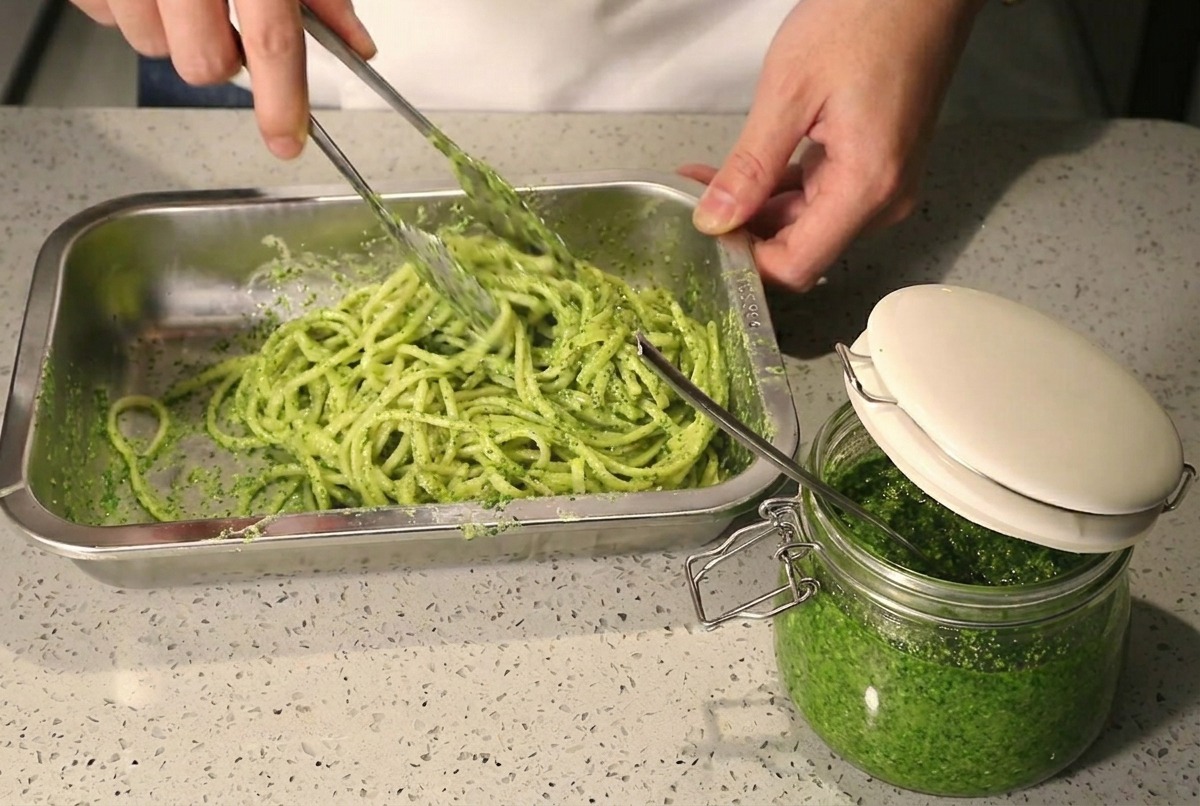 Using metal tongs to stir and mix spaghetti with green pesto and white cream in a metal tray.