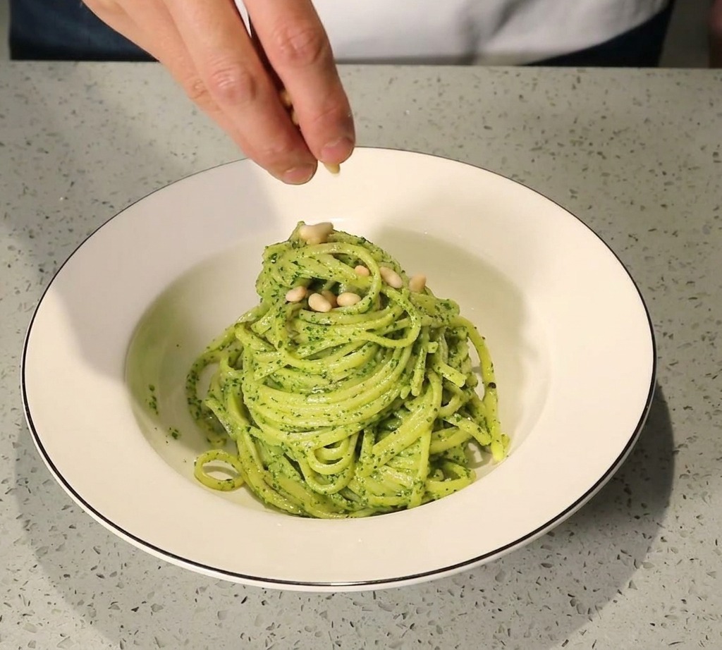 A hand sprinkling golden toasted pine nuts onto a neatly plated serving of pesto pasta in a white bowl.