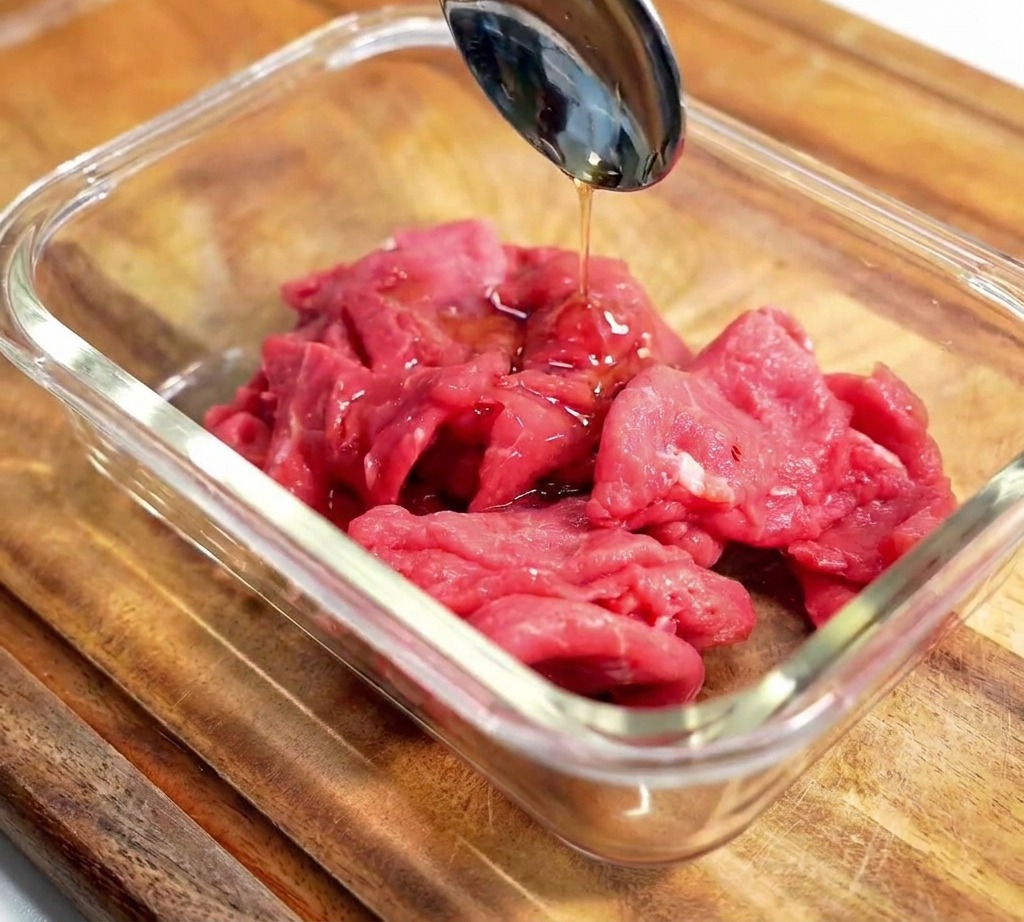 A metal spoon pouring dark liquid seasoning over sliced raw beef in a glass bowl on a wooden surface.