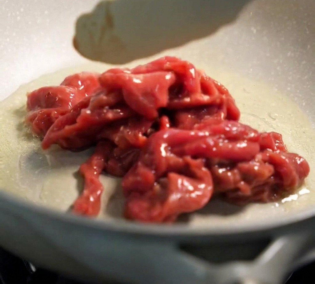 Tongs placing raw marinated beef into a hot, oiled frying pan.
