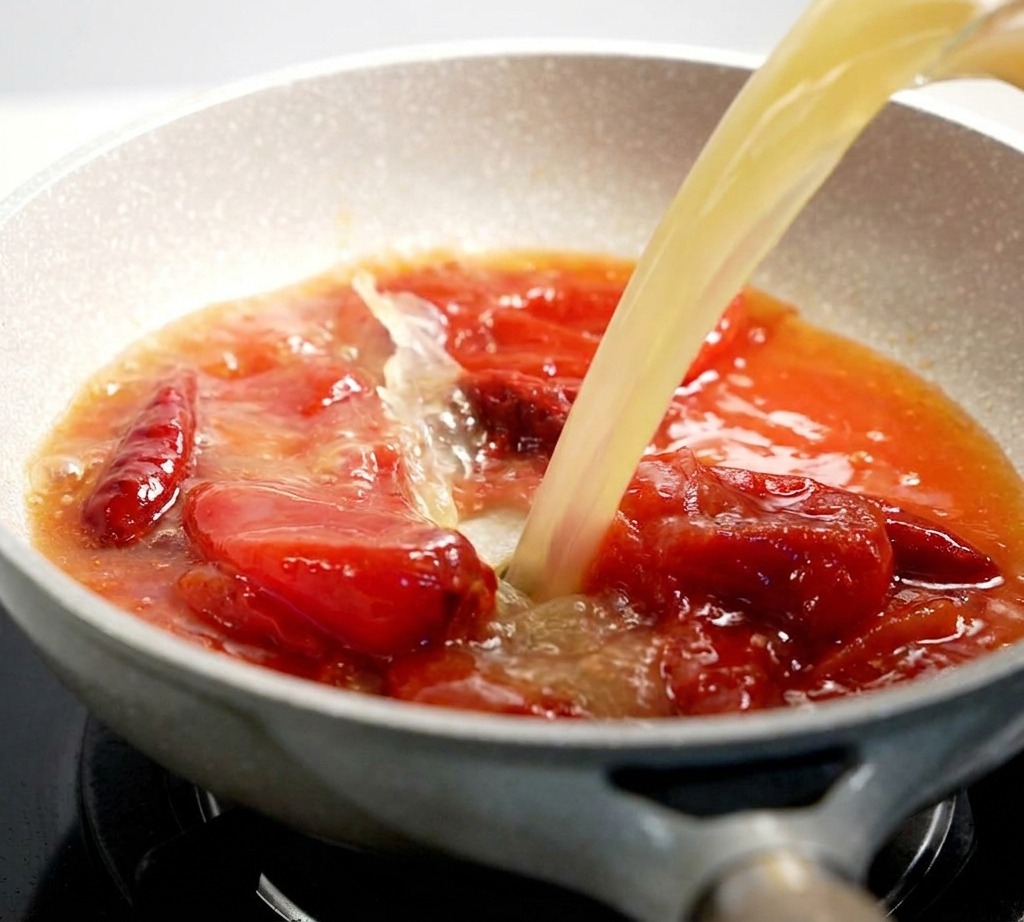 A light-colored broth being poured into a pan filled with a bubbling red tomato sauce mixture.