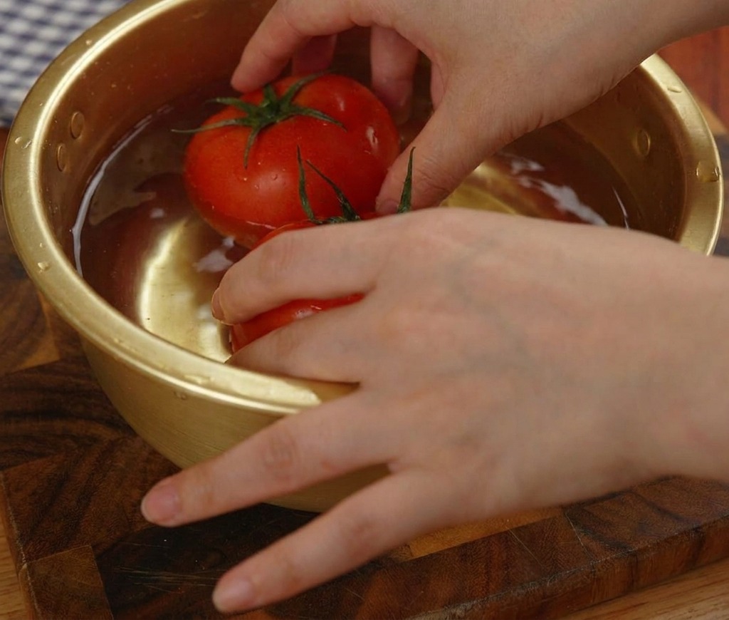 Two whole red frozen tomatoes being submerged and rotated in a golden metal bowl filled with water.