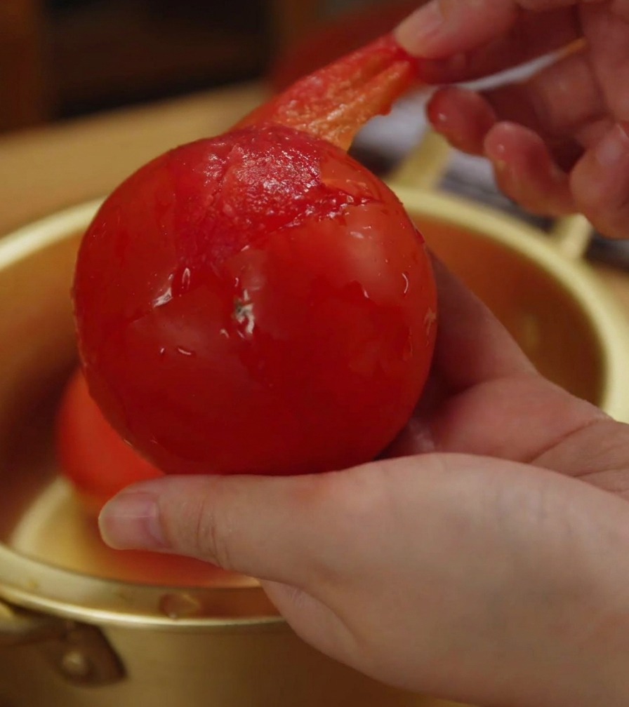 A close-up of a hand peeling the thin, translucent red skin off a whole, softened tomato.