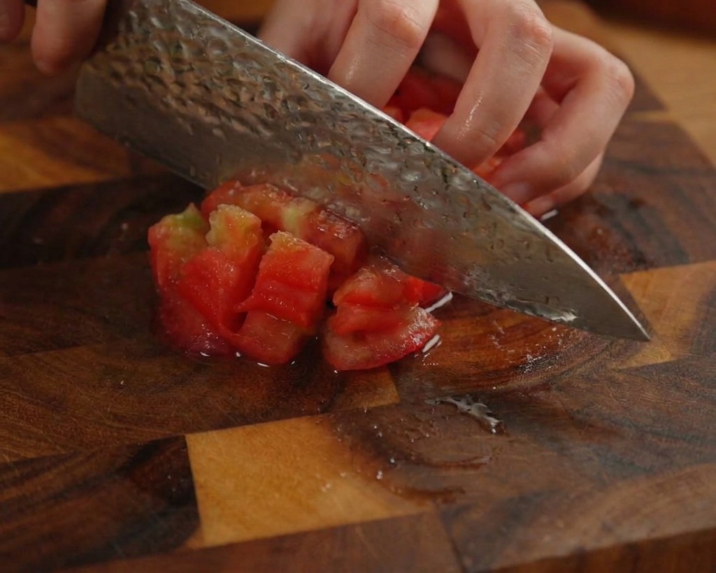 A person dicing a peeled red tomato into small chunks on a dark wooden board using a large kitchen knife.