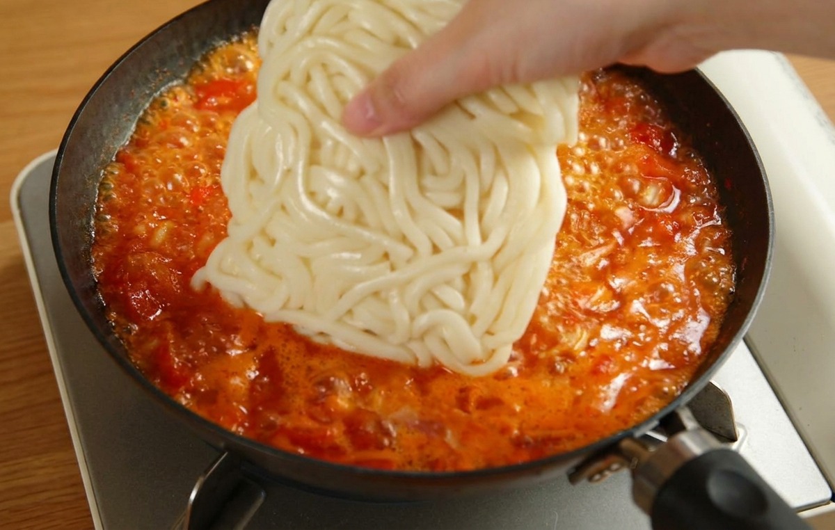A hand placing a square block of white udon noodles into a pan of simmering red tomato sauce.