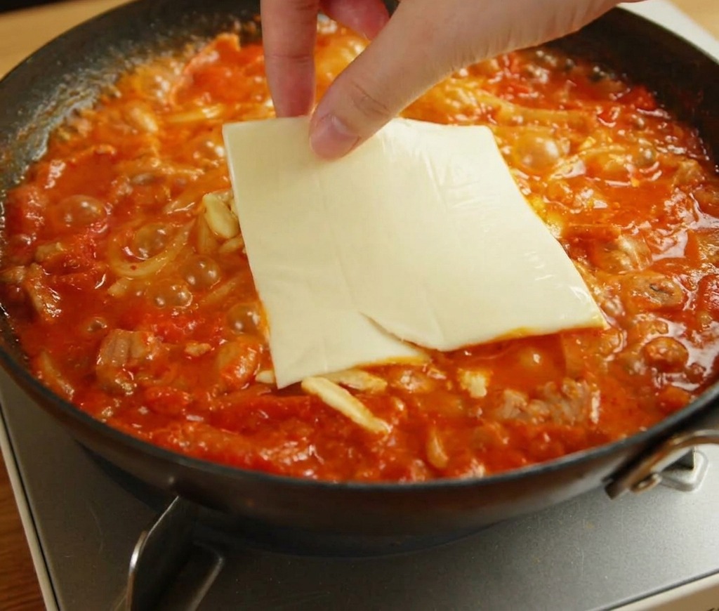 A hand placing a square slice of yellow American cheese on top of melting mozzarella and udon noodles.
