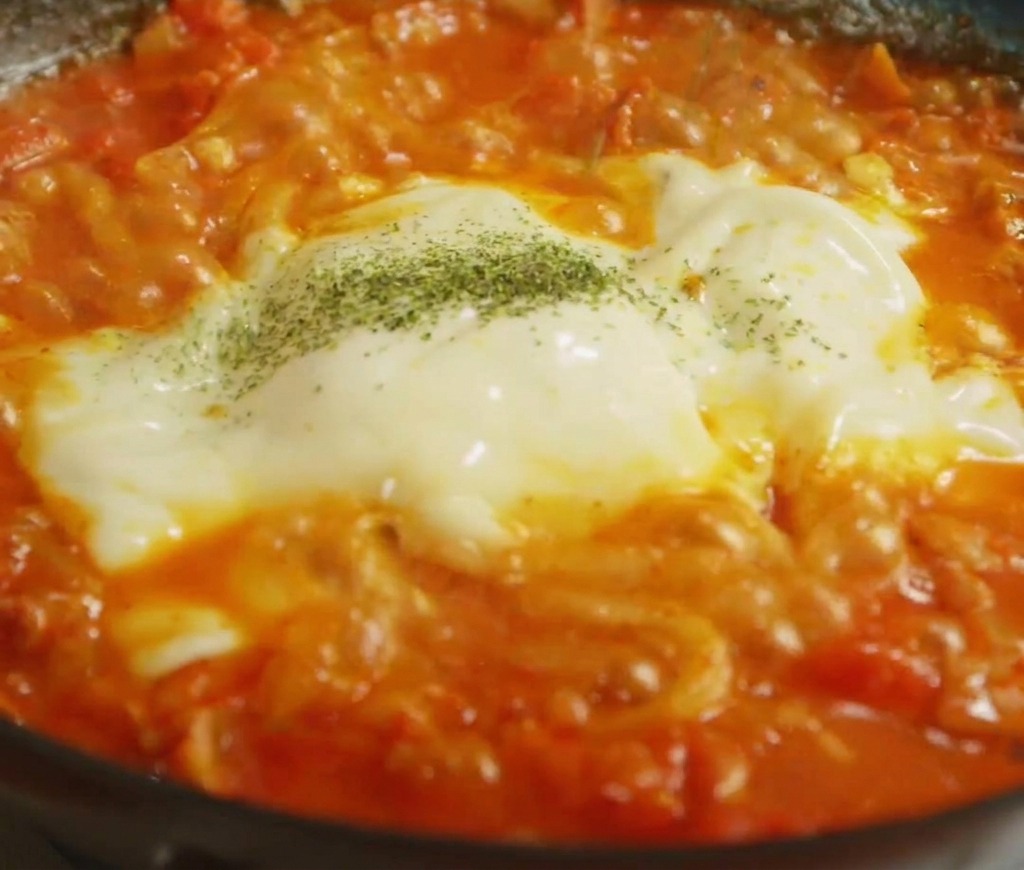 Dried green herbs being sprinkled over a thick layer of melted white and yellow cheese on top of noodles.