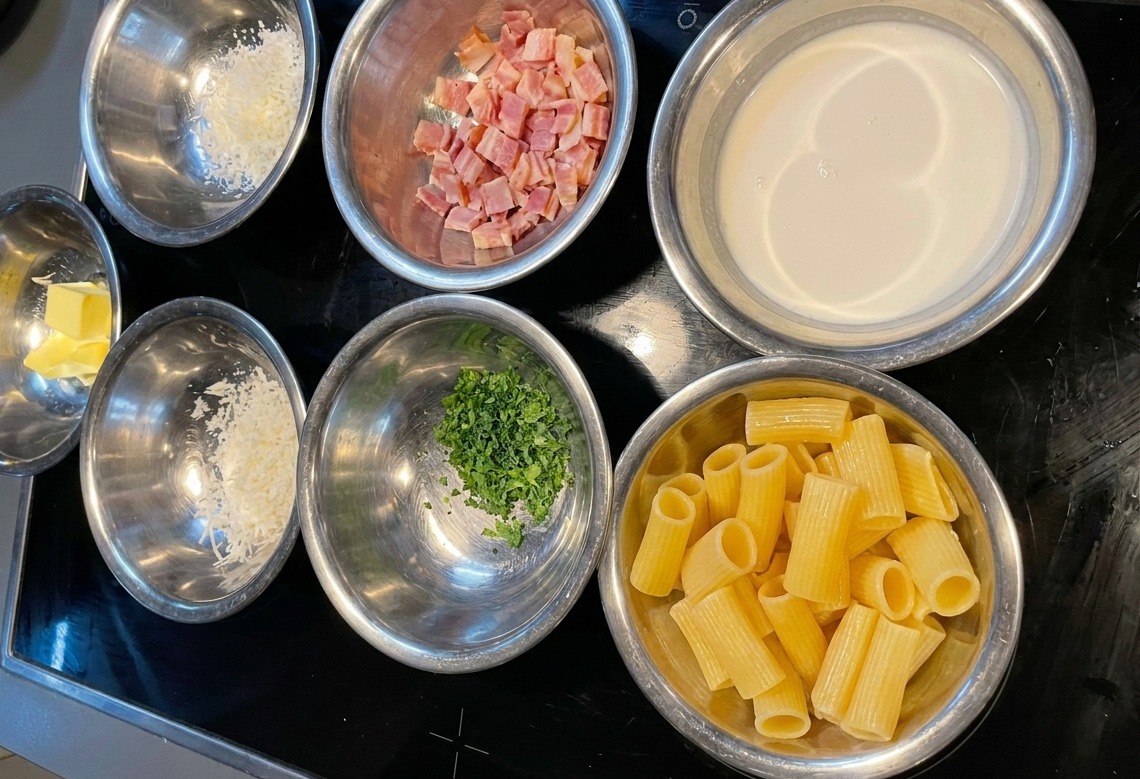 Several stainless steel bowls containing measured portions of rigatoni, heavy cream, chopped bacon, grated cheese, and parsley arranged on a black stovetop.