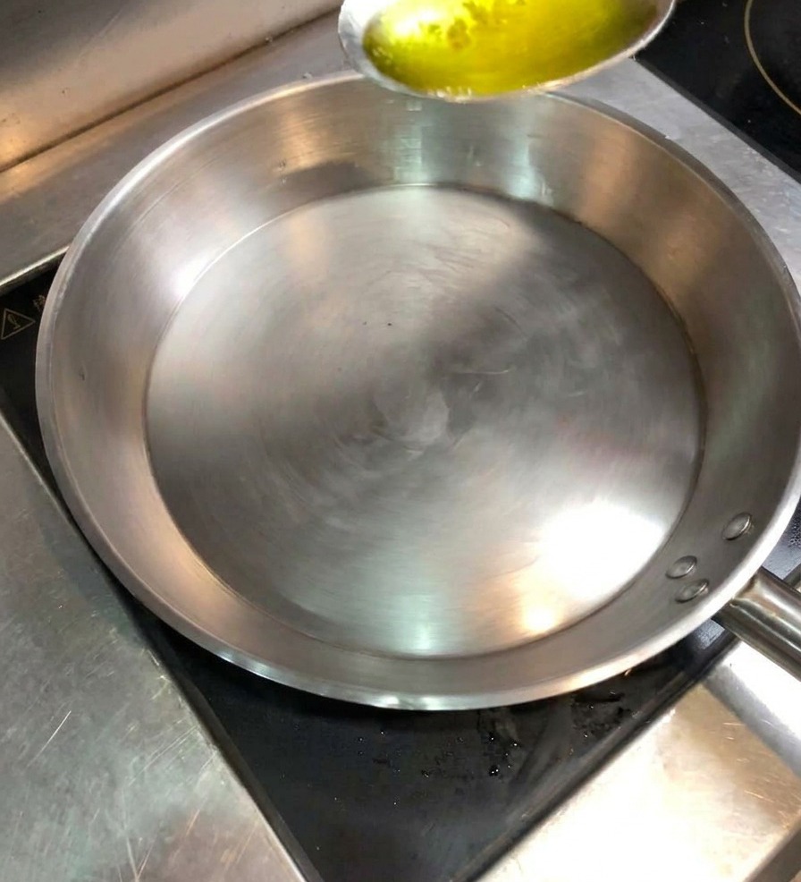 A metal spoon pouring golden cooking oil into a clean, heated stainless steel sauté pan.