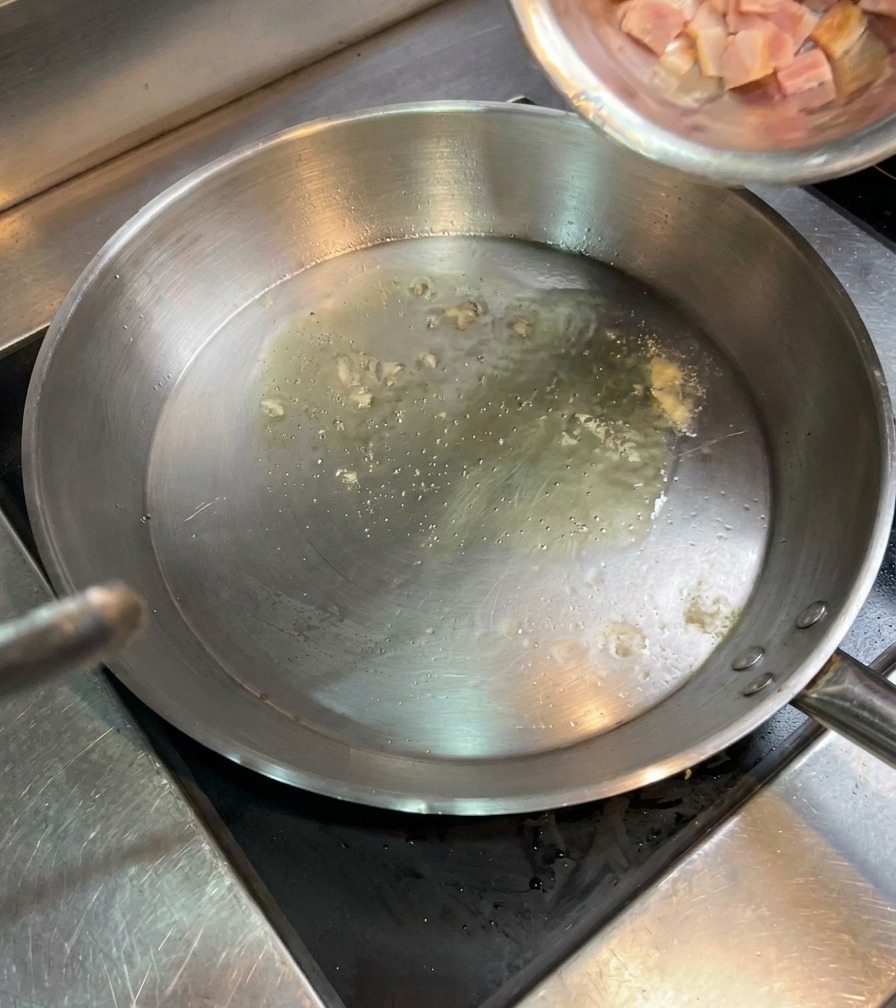 Small cubes of raw bacon being poured from a stainless steel bowl into a pan with shimmering hot oil.