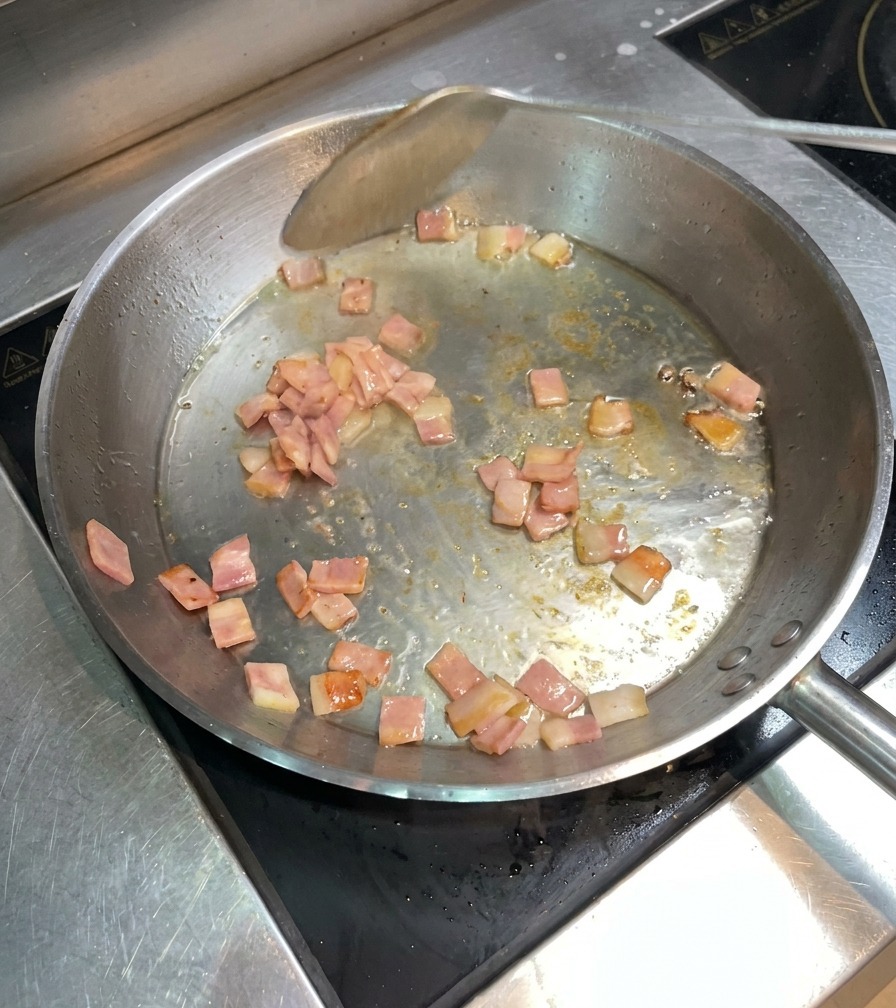 Chopped bacon pieces sizzling and browning in a pan, being moved around with a metal spatula.
