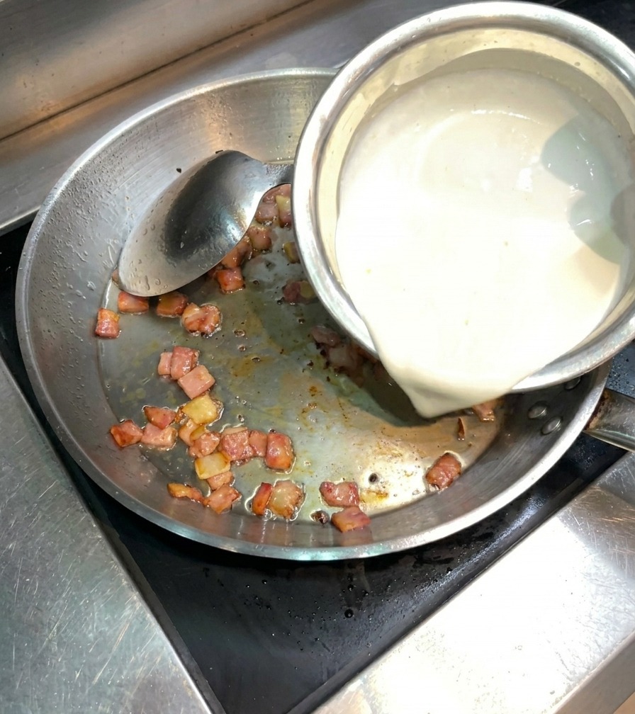 Thick white heavy cream being poured from a bowl into a stainless steel pan containing crispy sautéed bacon.