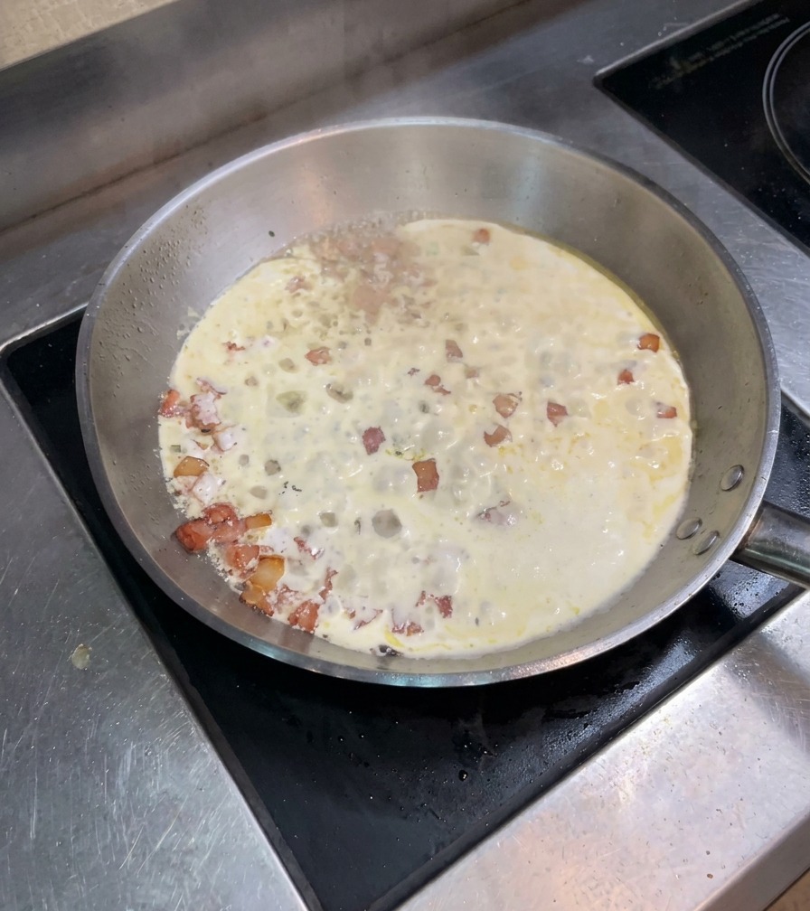 A mixture of heavy cream and bacon bits simmering and bubbling in a wide stainless steel pan on the stove.