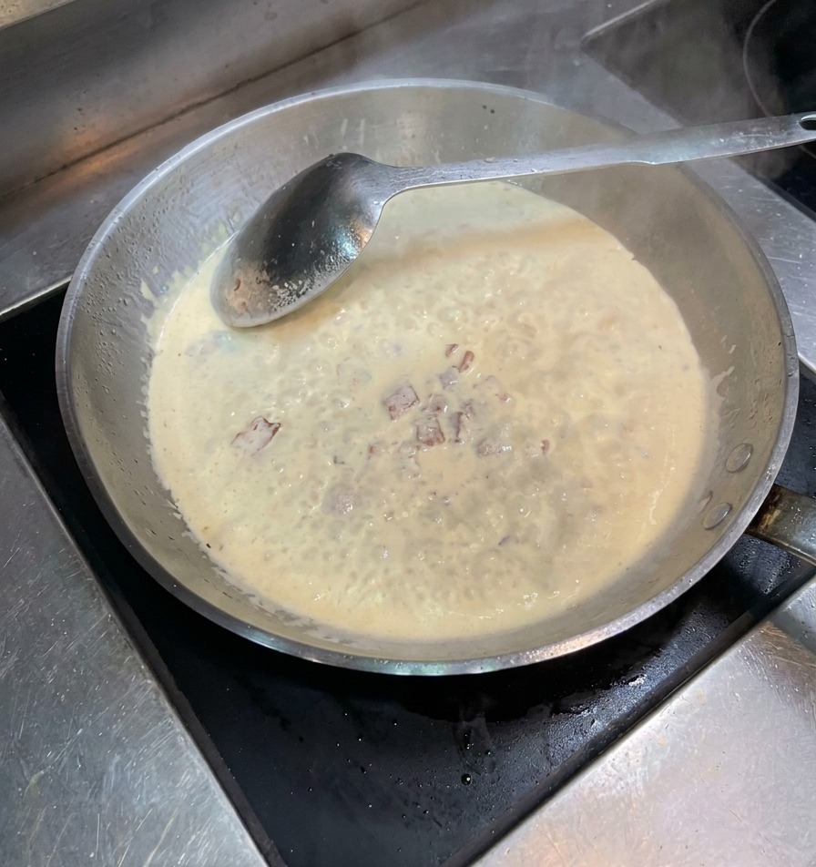 Black pepper and salt being sprinkled into a pan of simmering cream and bacon bits.