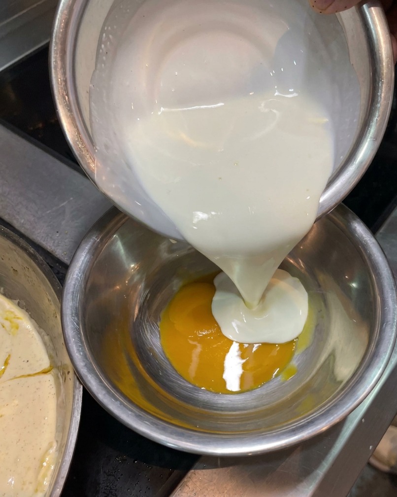 Heavy cream being poured from a silver bowl into another bowl containing a bright yellow egg yolk.
