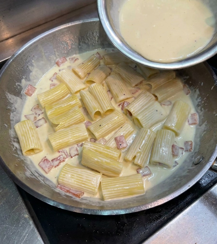 A creamy yellow egg and cheese mixture being poured over rigatoni pasta in a large metal pan.