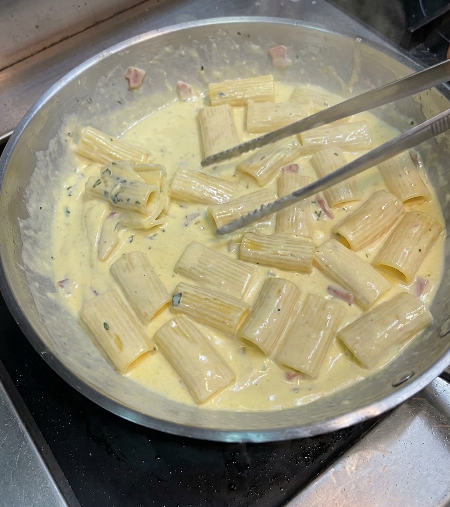 Tongs tossing rigatoni pasta in a pan to coat it thoroughly in a thick, creamy carbonara sauce.