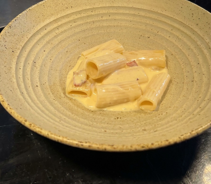 A portion of rigatoni coated in a glossy, pale yellow cream sauce with visible bacon bits, served in a wide, textured ceramic bowl.