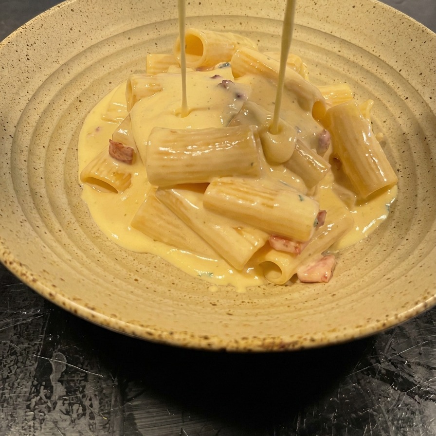 Thick, velvety cream sauce being poured from a pan over a mound of rigatoni pasta in a speckled ceramic bowl.