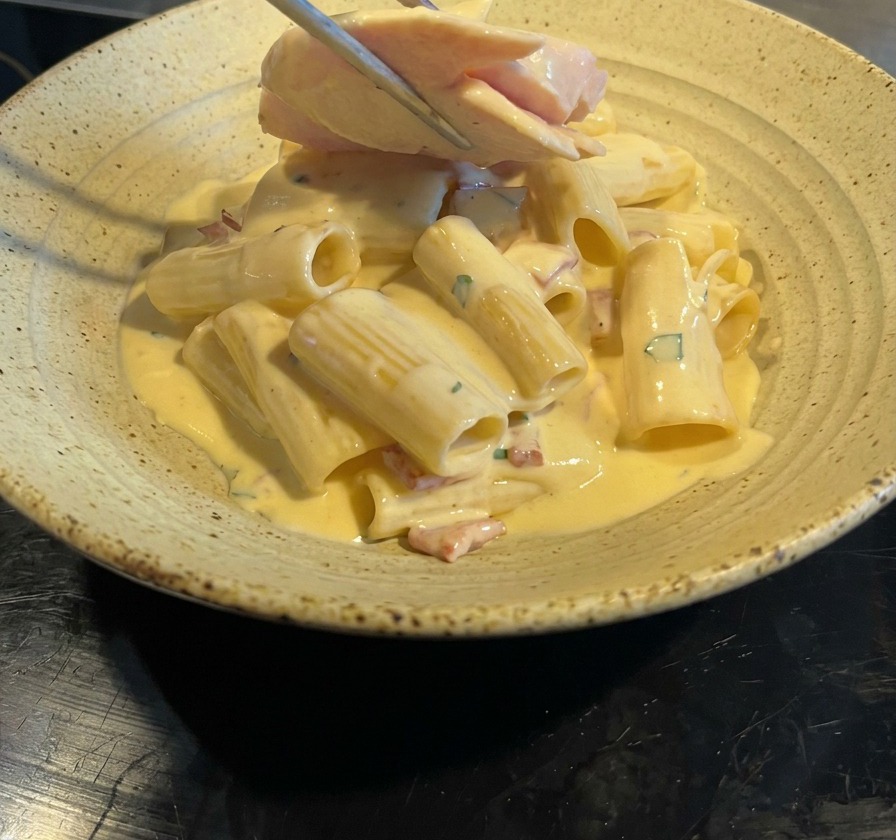 Metal tongs placing several slices of pan-seared chicken breast on top of a serving of creamy bacon rigatoni.