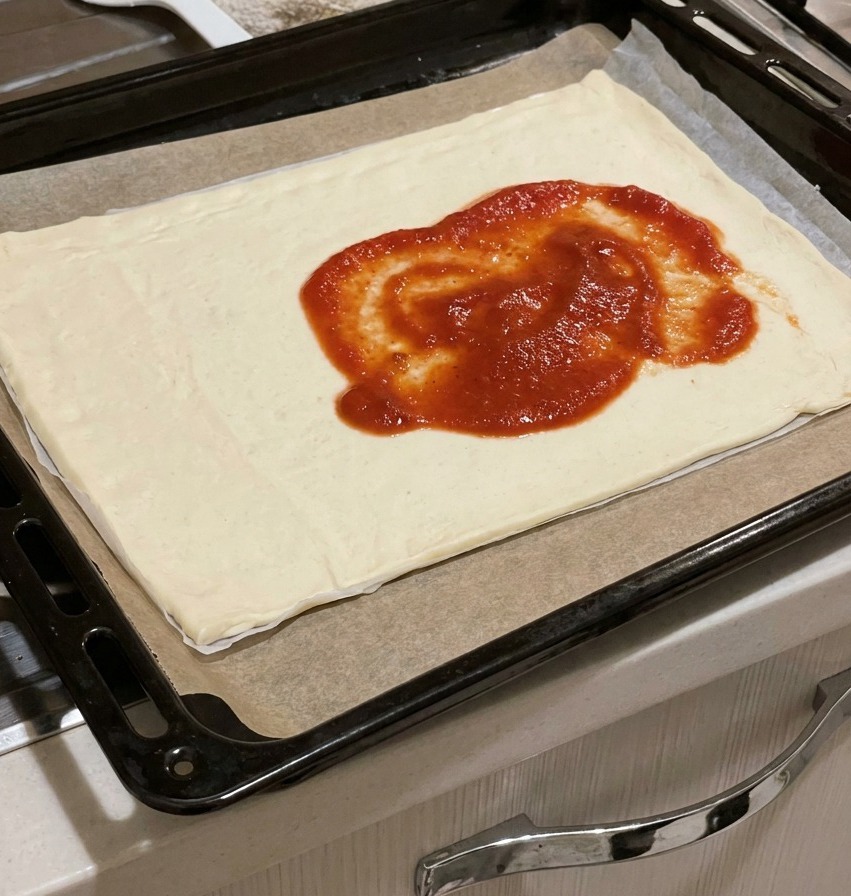 Thick red tomato sauce dolloped in the middle of a raw rectangular pizza dough on a tray.